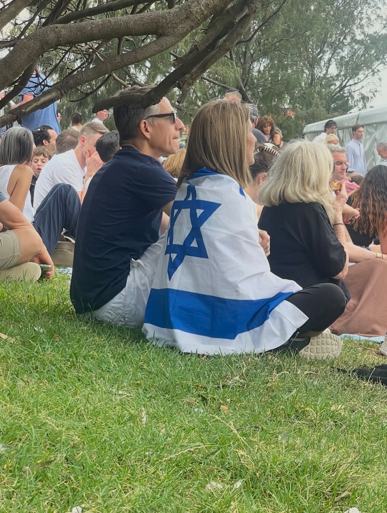 Person wearing an Israeli flag shirt sitting on grass with a crowd in the background