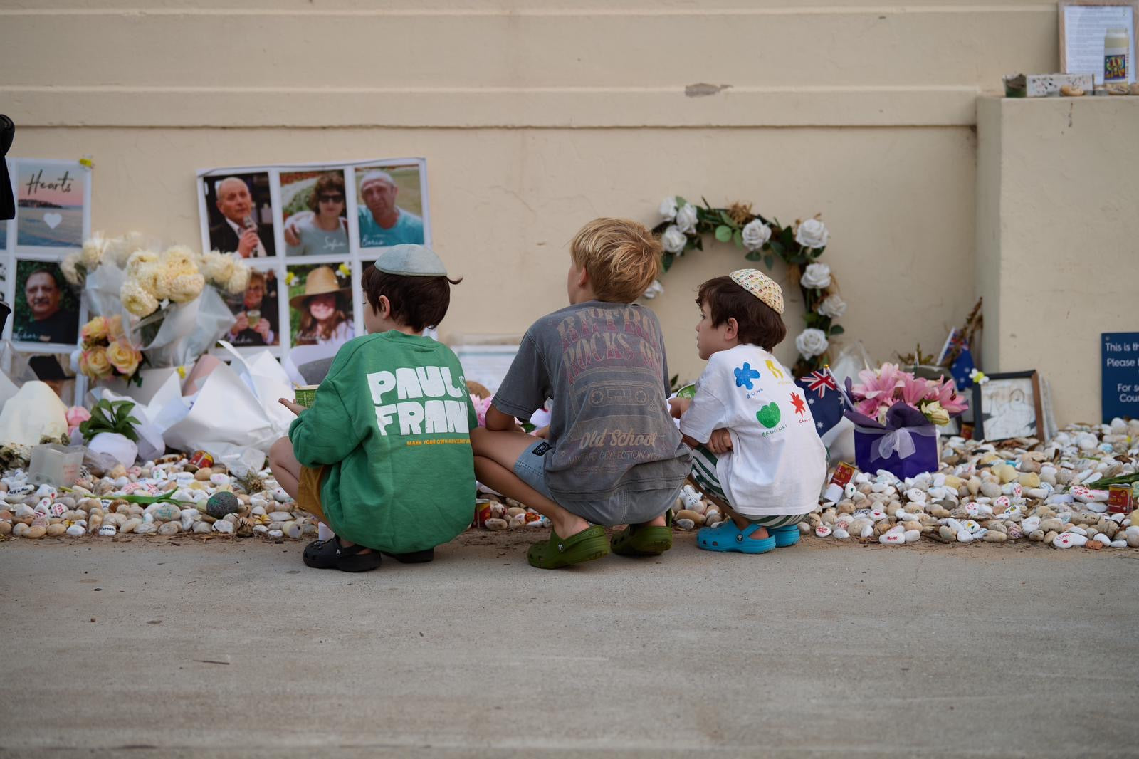 Three children sitting on the ground in front of a memorial with flowers, photos, and a wreath.