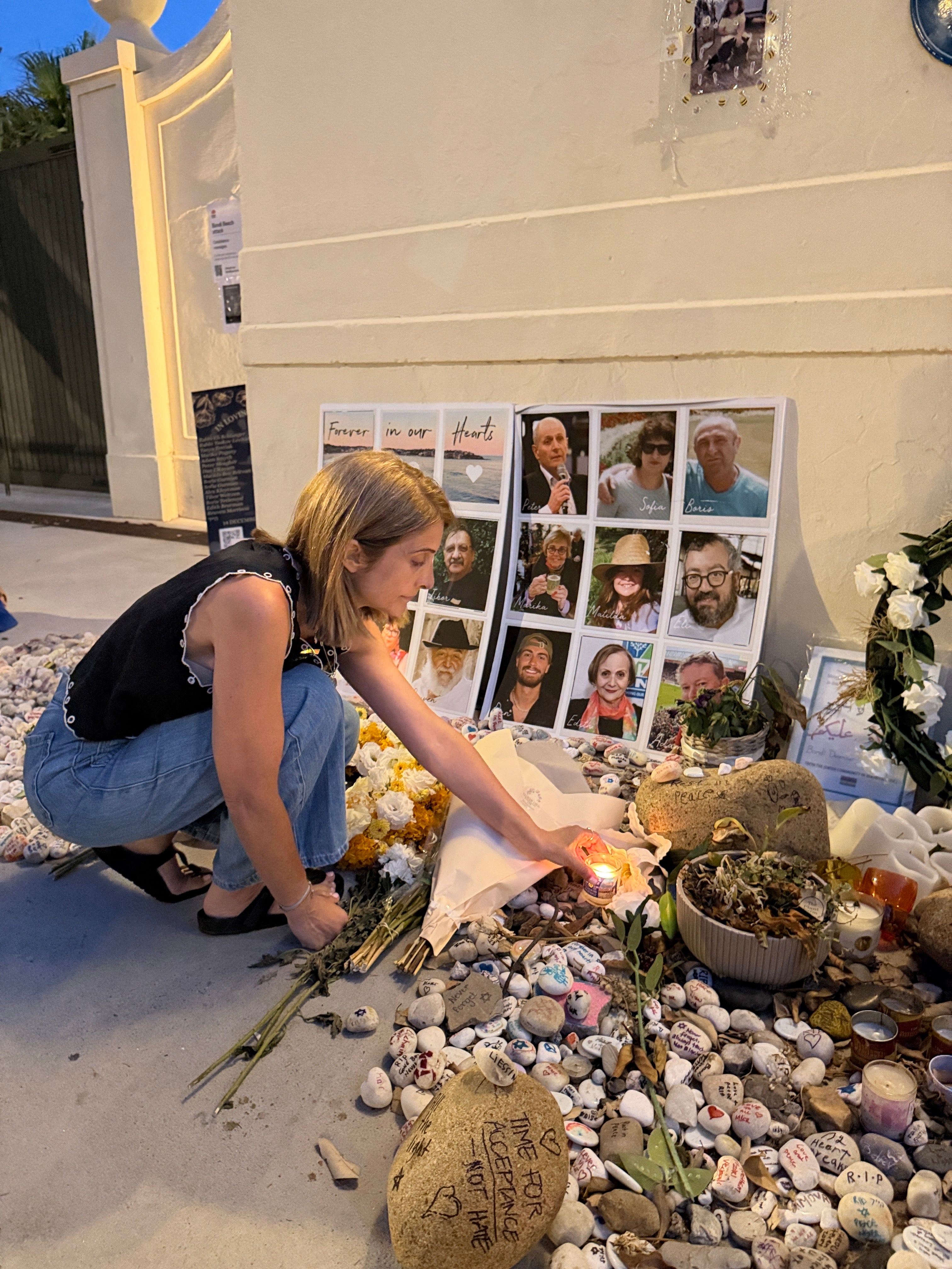Person lighting a candle at a memorial with photos and offerings on a stone wall.