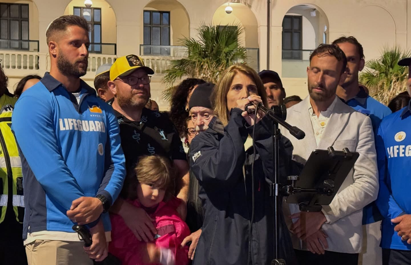 Group of people, including a person in a blue 'Lifeguard' shirt, gathered outdoors with a microphone and camera.